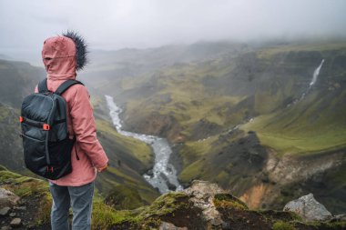 Leylak rengi ceketli ve sırt çantalı bir kadın İzlanda 'da Haifoss şelalesi yakınlarında İzlanda dağ ve nehir çukurlarının keyfini çıkarıyor..