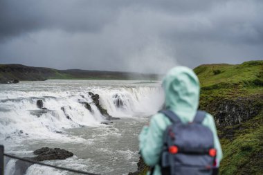 Sırt çantalı ve yeşil ceketli kadın İzlanda 'da Gullfoss' un güçlü şelalesinin önünde duruyor. İzlanda 'nın Altın Çember rotasını takip eden turistik ilgi odağı.