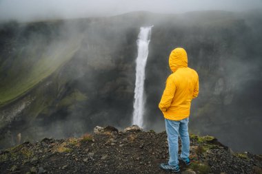 Yağmurlu bulutlu havada Haifoss şelalesinin keyfini çıkaran sarı ceketli bir adam. İzlanda 'nın İskoçya' sı.