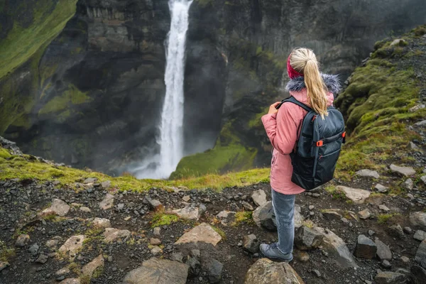 Woman with backpack and lilac jacket enjoying Haifoss waterfall of Iceland Highlands in Thjorsardalur Valley.