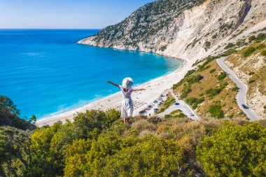 Mutlu bir kadın bir kayanın üzerinde duruyor, heyecan verici bir özgürlük duygusuyla el kaldırıyor, Myrtos Sahili 'ne bakıyor. Sefalonya Adası, Yunanistan.
