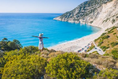 Mutlu bir kadın bir kayanın üzerinde duruyor, heyecan verici bir özgürlük duygusuyla el kaldırıyor, Myrtos Sahili 'ne bakıyor. Sefalonya Adası, Yunanistan.