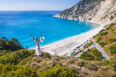 Mutlu bir kadın bir kayanın üzerinde duruyor, heyecan verici bir özgürlük duygusuyla el kaldırıyor, Myrtos Sahili 'ne bakıyor. Sefalonya Adası, Yunanistan.