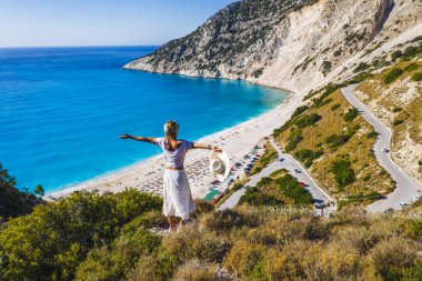 Mutlu bir kadın bir kayanın üzerinde duruyor, heyecan verici bir özgürlük duygusuyla el kaldırıyor, Myrtos Sahili 'ne bakıyor. Sefalonya Adası, Yunanistan.
