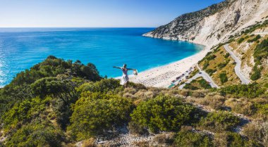 Mutlu bir kadın bir kayanın üzerinde duruyor, heyecan verici bir özgürlük duygusuyla el kaldırıyor, Myrtos Sahili 'ne bakıyor. Sefalonya Adası, Yunanistan.