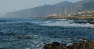 Coastline with houses, Los Gigantes cliffs in Puerto de Santiago, Santiago del Teide, Tenerife. Big waves crash on the rocky shore.