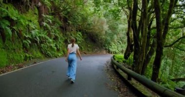 Woman walks along an asphalt road in a dense forest. Overgrown trees in laurel tree forest or jungle, Anaga National Park, Tenerife, Canary Islands.