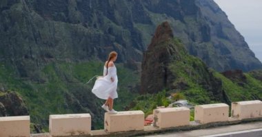 Elegant woman enjoying tropical nature view. Inspired freedom girl admiring landscape of majestic Masca gorge valley. Tenerife, Spain. Blonde woman with a long hair braid, dressed in a white dress.