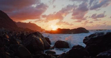 Foaming white sea water sprays over the rugged black rock coast in Tenerife at sunset. Cinematic shot of ocean waves crashing into the volcanic shore of Canary island at golden hour