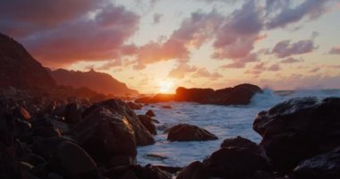 Rock formations on Tenerife beach during sunset. Static shot. Atlantic ocean. Orange clouds and silhouette of mountains, waves splashing. Meditation