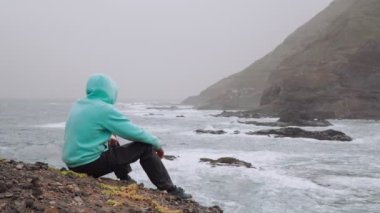 Güçlü dalgaların önünde elinde harita olan gezgin kayalık kıyı şeridine sıçrıyor. Yürüyüş yolunda engebeli volkanik dağlar. Santo Antao Adası, Cape Verde. 4K görüntü