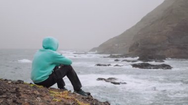 Traveler enjoy the powerful waves splashing into rocky coastline. Huge volcanic mountains rugged on the trekking path. Santo Antao Island, Cape Verde. 4K footage.