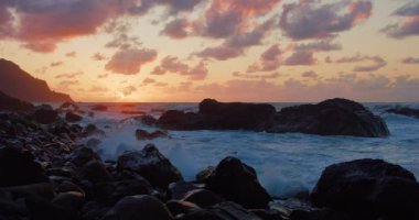 Rock formations on Tenerife beach during sunset. Static shot. Atlantic ocean. Orange clouds and silhouette of mountains, waves splashing. Meditation