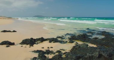 View of a white sand beach with black rocks. Blue transparent ocean waves splashing foam. Shoreline of paradise tourist destination. Relax travel concept. Nobody.