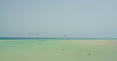 Colorful kite shape flying on blue clear sky. Many people ride on the waves on the weekend. Shallow transparent ocean waters. Fuerteventura, Grandes Playas Corralejo. Canary Islands.