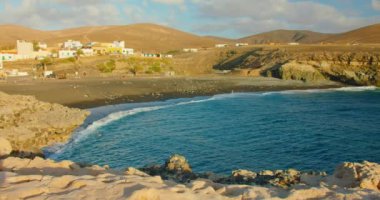 Spain, Canary Islands, Fuerteventura, sandy volcanic beach in little town Ajuy. Blue ocean waves crash on black seashore. Cityscape with resort buildings. Playa de Ajuy.