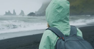 Yeşil yağmurluklu bir kadın kara kumları ziyaret ediyor. Reynisfjara 'nın deniz kenarındaki uçurum oluşumlarına bakıyor..