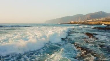 Fırtınalı havada insansız hava aracı okyanus yüzeyinde alçaktan uçuyor. Deniz dalgaları köpürüyor. Kaya kıyısı. Arka planda Los Gigantes uçurumları var. Tenerife. Turkuaz Atlantik sularının güzel manzarası. Kanarya