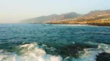 Fırtınalı havada insansız hava aracı okyanus yüzeyinde alçaktan uçuyor. Deniz dalgaları köpürüyor. Kaya kıyısı. Arka planda Los Gigantes uçurumları var. Tenerife. Turkuaz Atlantik sularının güzel manzarası. Kanarya