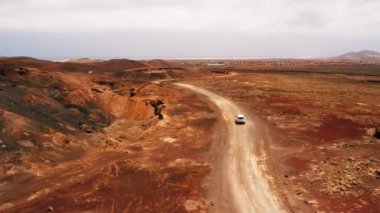 Tek başına giden beyaz bir SUV, güneşli yaz gününde çöldeki volkanik arazide boş toprak yolda seyahat ediyor. Hava aracının geniş görüşü. Araç ülke yolunda ilerliyor. Fuerteventura