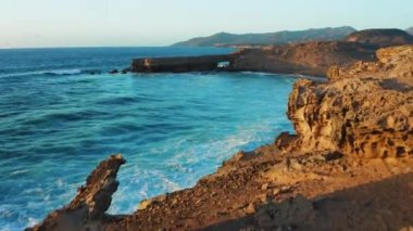 Gün batımında mavi Atlantik Okyanusu suyuyla birlikte kayalık sahil şeridinde. Doğa manzarası. Altın kumtaşı blöf plajı. Seyahat güzergahı. Fuerteventura Adası, Kanaryalar. 4K sinematik görünüm