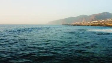 Fırtınalı havada insansız hava aracı okyanus yüzeyinde alçaktan uçuyor. Deniz dalgaları köpürüyor. Kaya kıyısı. Arka planda Los Gigantes uçurumları var. Tenerife. Turkuaz Atlantik sularının güzel manzarası. Kanarya