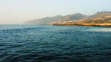 Fırtınalı havada insansız hava aracı okyanus yüzeyinde alçaktan uçuyor. Deniz dalgaları köpürüyor. Kaya kıyısı. Arka planda Los Gigantes uçurumları var. Tenerife. Turkuaz Atlantik sularının güzel manzarası. Kanarya