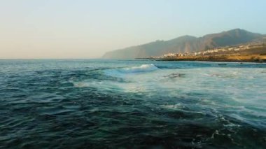 Fırtınalı havada insansız hava aracı okyanus yüzeyinde alçaktan uçuyor. Deniz dalgaları köpürüyor. Kaya kıyısı. Arka planda Los Gigantes uçurumları var. Tenerife. Turkuaz Atlantik sularının güzel manzarası. Kanarya