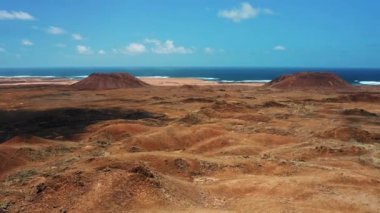 Las Calderas, Caldera Encantada 'da terk edilmiş boş topraklarda yürüyüş alanı. Çorak kahverengi topraklar üzerinde iki volkan yükseliyor. Mavi gökyüzü. Dev volkanik kraterlerin üzerinde uçan, gök mavisi okyanuslar. Fuerteventura. Kanarya