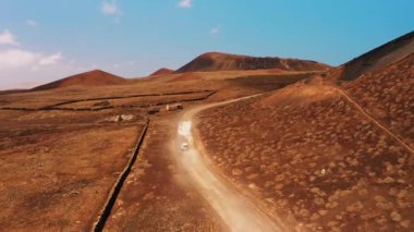 Kızıl çöl manzarasında dar bir yolda ilerleyen beyaz arabayı takip eden hava görüntüsü. Vechicle toprak yolda hızla tozlanarak ilerliyor. Her yere git konsepti. Seyahat zamanı. Geniş hava aracı