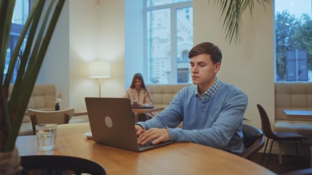 セーターを着た若者がカフェに座り ノートパソコンに入力すると 背景がぼやけている女性がスマートフォンを使っている アークはフリーランサーを遠隔操作で撃った 自己雇用の概念 — ストック動画