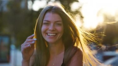 Güzel bir kadın kameraya gülüyor ve gün batımında uzun saçlarına dokunuyor. Arka planda bulanık bir sokak. Dışarıdaki mutlu kadın modelin portresi. Duygu kavramı