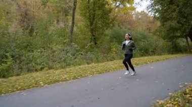 Spor kıyafetli, kulaklıklı, parkta koşu yapan genç bir kadın. Serin bir sabahta aktif kadın eğitimi. Sağlıklı yaşam tarzı kavramı
