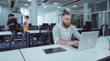 Açık uzay ofisinde dizüstü bilgisayarda çalışan kızıl saçlı sakallı bir adam yanında tablet olan, arka planda bulanık konuşan iş arkadaşları. Ark çalışanı internet kullanarak vurdu. İş kavramı