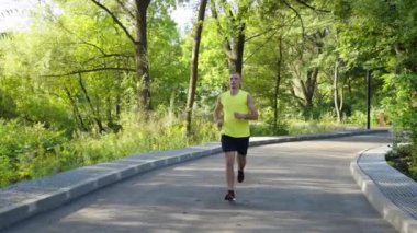 Yavaş çekim atletik adam, sarı gömlek ve siyah şort giymiş, güneşli bir sabahta parkta koşuyor. Dışarıdaki koşucu eğitiminin izini sürüyorum. Sağlıklı yaşam tarzı kavramı