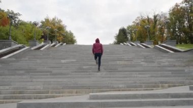 Spor giyimli, ağır çekimde, kafasında kapüşonlu, parktaki merdivenlerde koşan, yanında sarı ağaçlar olan adam. Atış talimini takiben erkek sporcu, sonbahar sabahı dışarıda. Zindelik kavramı