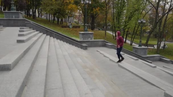 穿着运动服 头戴头罩的慢动作男子在城市公园宽阔的楼梯上跑来跑去 背景是黄色的树 秋季早上追踪射击慢跑者的运动 健康的概念 — 图库视频影像