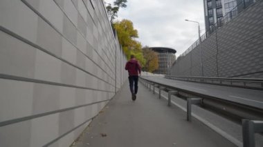 Spor kıyafetleri içinde ağır çekim formundaki bir adam bir tarafta yüksek duvarın yanında ve diğer tarafta yol kıvrımlı, uzaktaki binalar, duvarın üzerinde sarı ağaçlar. Koşucunun sonraki çekimi