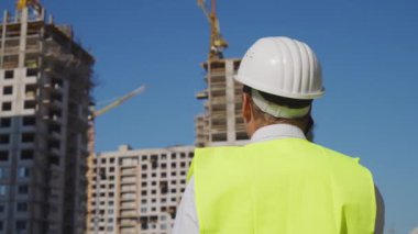 İnşaat alanında güvenlik yeleği ve şapka takan bir mühendis telefonla konuşuyor. Güneşli bir günde arka planda çalışan adam. Sanayi kavramı