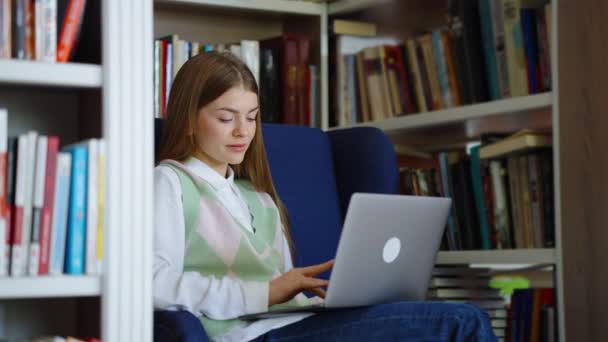 本棚の間のアームチェアに座ってノートパソコンを入力する学生 追跡は図書館で勉強するために若い女性のブラウジングインターネットを撃った 教育の概念 — ストック動画
