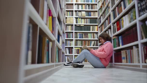 Афроамериканська дівчинка - підліток читає книжки в бібліотеці — стокове відео
