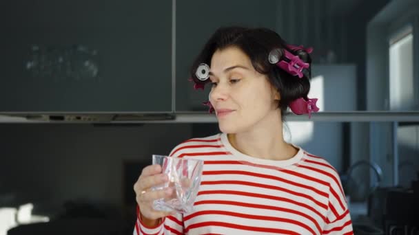 Женщина с бигуди для волос пьет воду на кухне — стоковое видео
