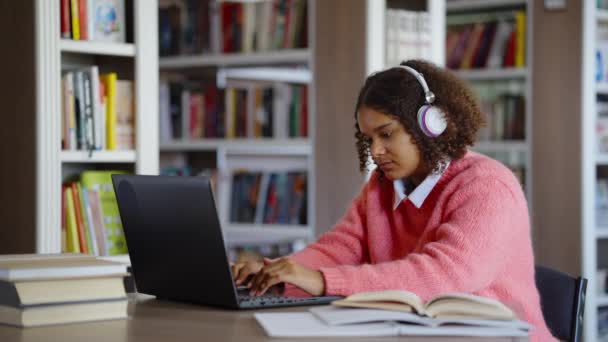 Czarna dziewczyna studiuje w bibliotece i przegląda internet na laptopie — Wideo stockowe