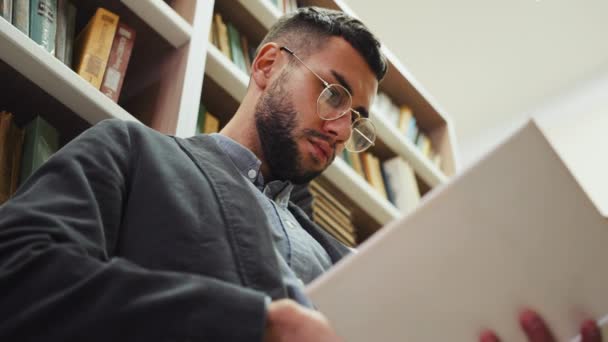 Чоловік в окулярах читає книгу в бібліотеці — стокове відео
