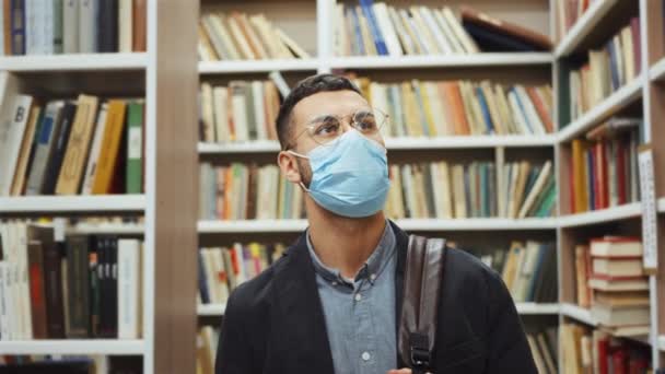 Studente in maschera che cammina tra le librerie in biblioteca — Video Stock