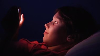 Çocuk gece yatağında akıllı telefondan video izliyor.