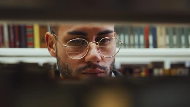 Silmälasipäinen mies kirjastossa kirjahyllyjen kautta katsottuna — kuvapankkivideo
