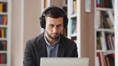 Gözlüklü adam kütüphanede kulaklık takıyor ve laptop kullanıyor.