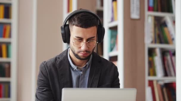 Mannen med glasögon som bär hörlurar och använder laptop i biblioteket — Stockvideo