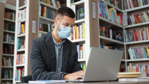 Muž v masce dělá online výzkum v knihovně — Stock video